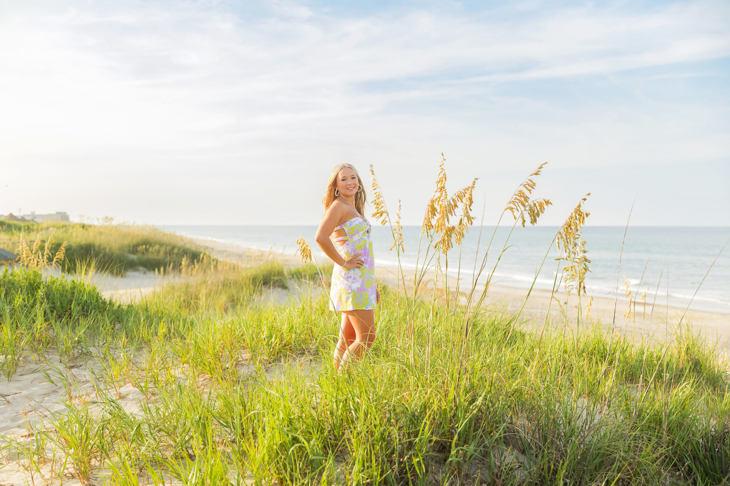 Outer Banks senior Annabelle in a pastel floral sundress standing in the sand dunes at Nags Head, smiling with beach grass and golden sunlight.