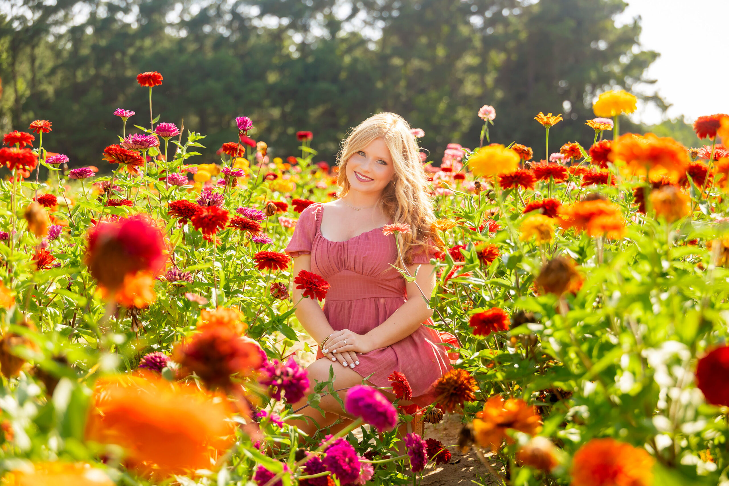 Senior girl standing in a wildflower field wearing a pink dress, surrounded by soft pink and green blooms in natural light.