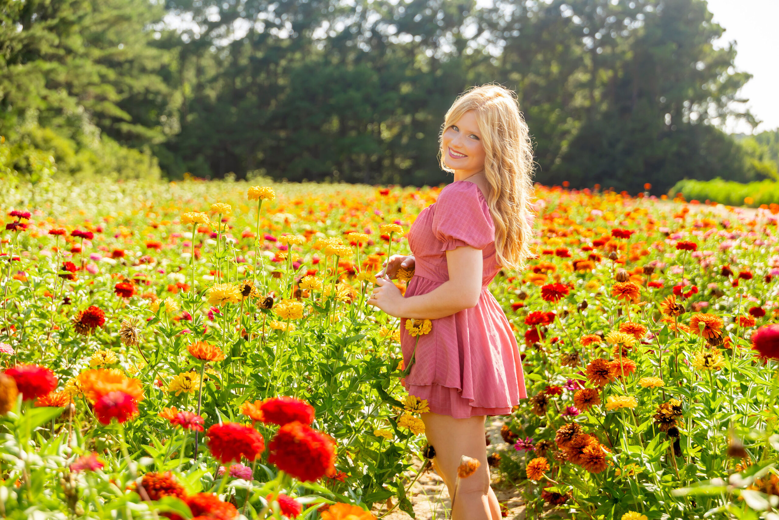 Senior girl standing in a wildflower field wearing a pink dress, surrounded by soft pink and green blooms in natural light.