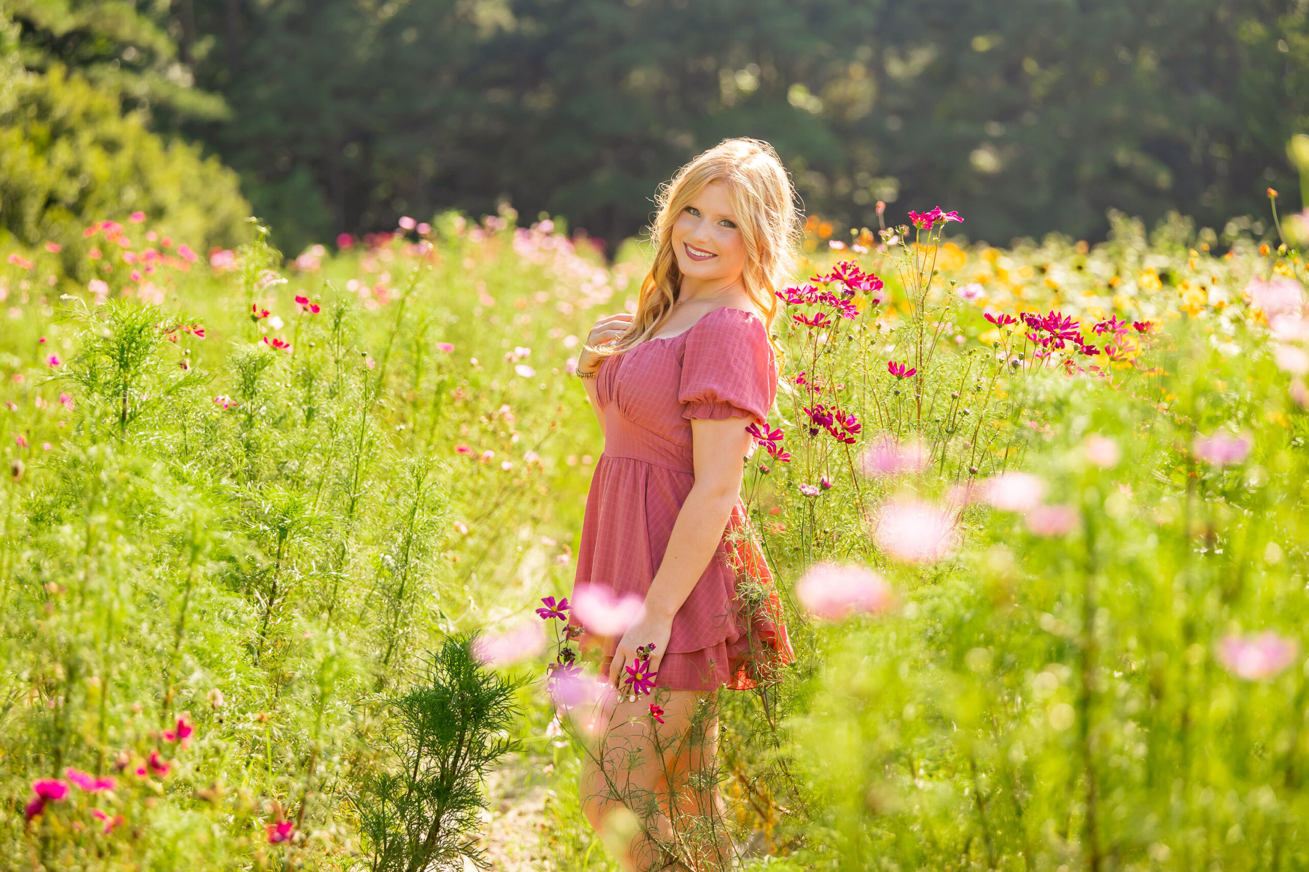 Senior girl standing in a wildflower field wearing a pink dress, surrounded by soft pink and green blooms in natural light.