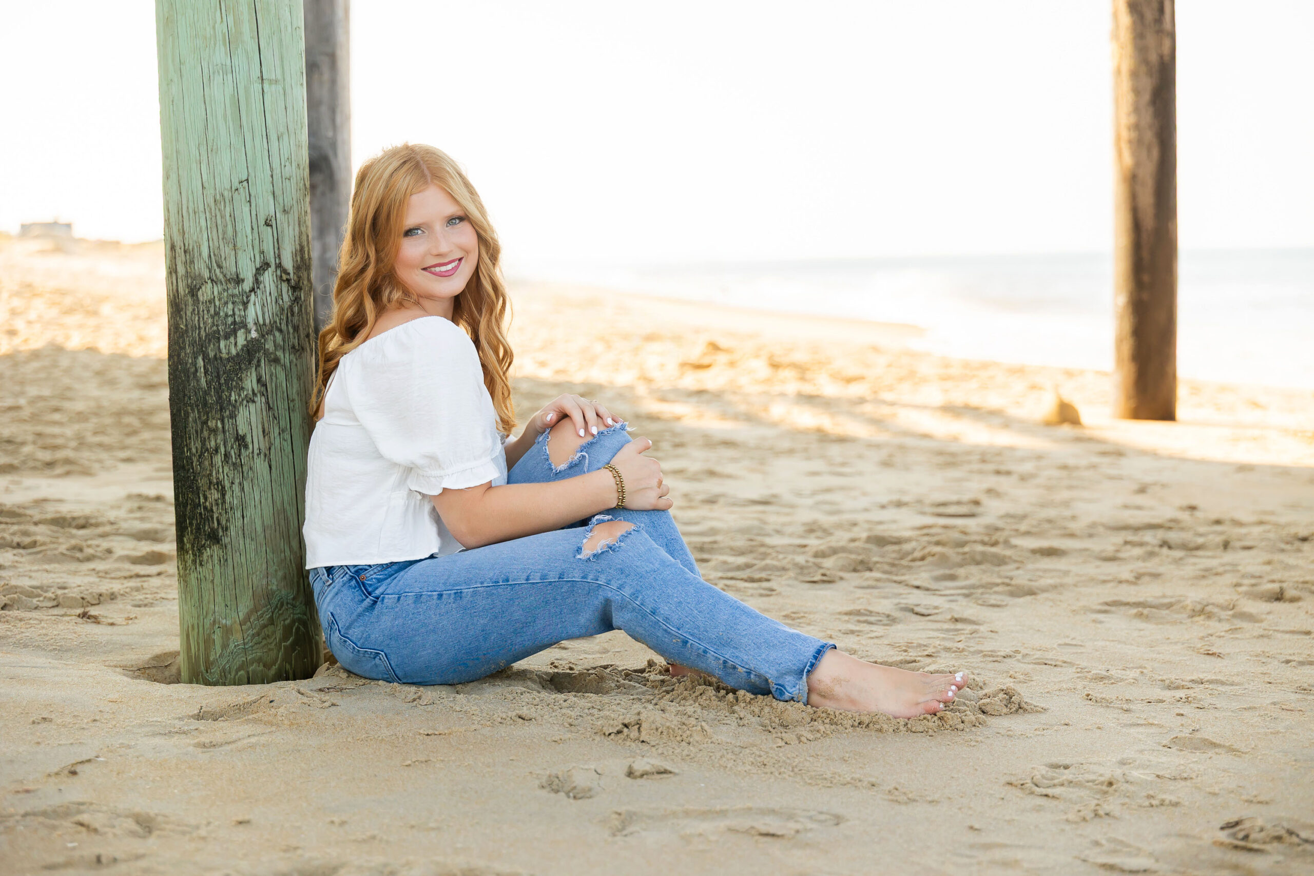 Senior girl sitting barefoot in the sand under the Kitty Hawk Fishing Pier wearing a white top and ripped jeans.