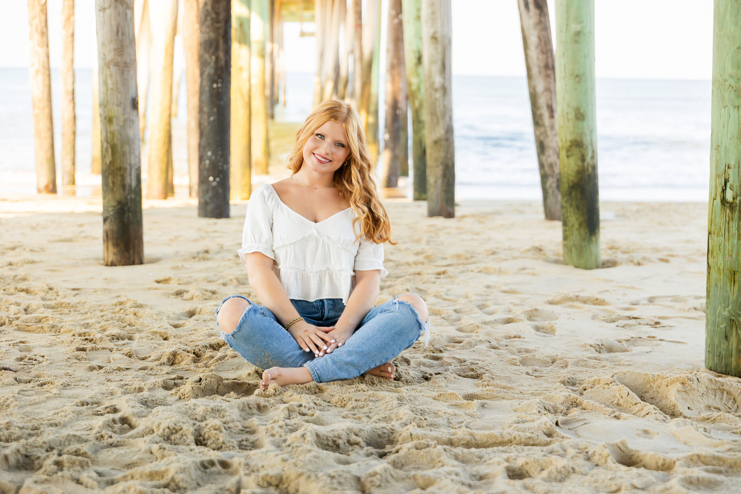 Senior girl sitting barefoot in the sand under the Kitty Hawk Fishing Pier wearing a white top and ripped jeans.