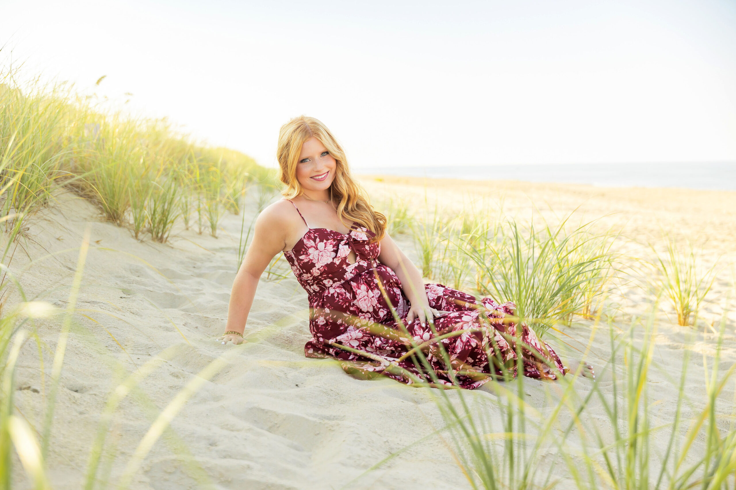 Senior girl standing in the beach dunes wearing a maroon floral maxi dress with sea grass and coastline behind her.