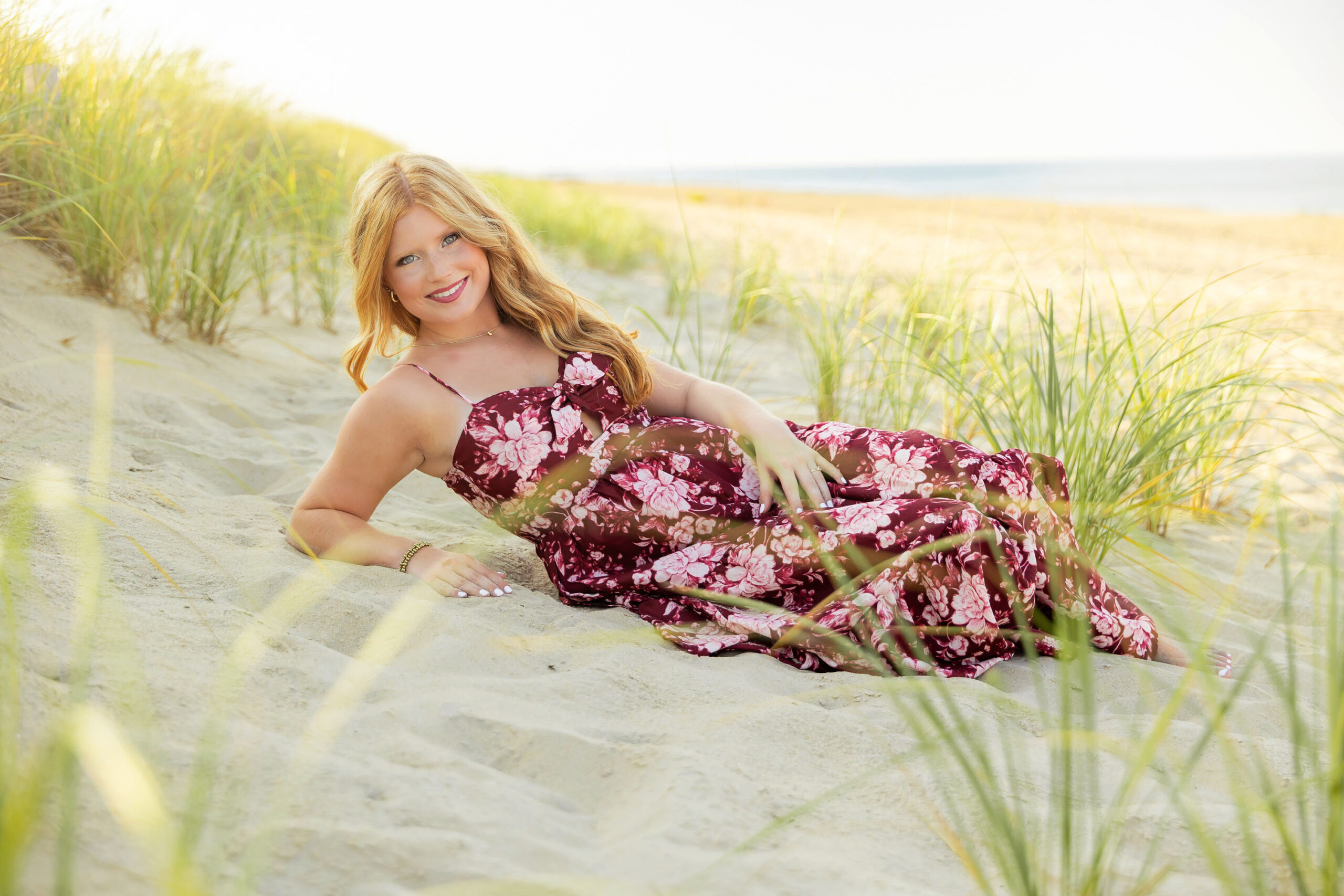 Senior girl standing in the beach dunes wearing a maroon floral maxi dress with sea grass and coastline behind her.