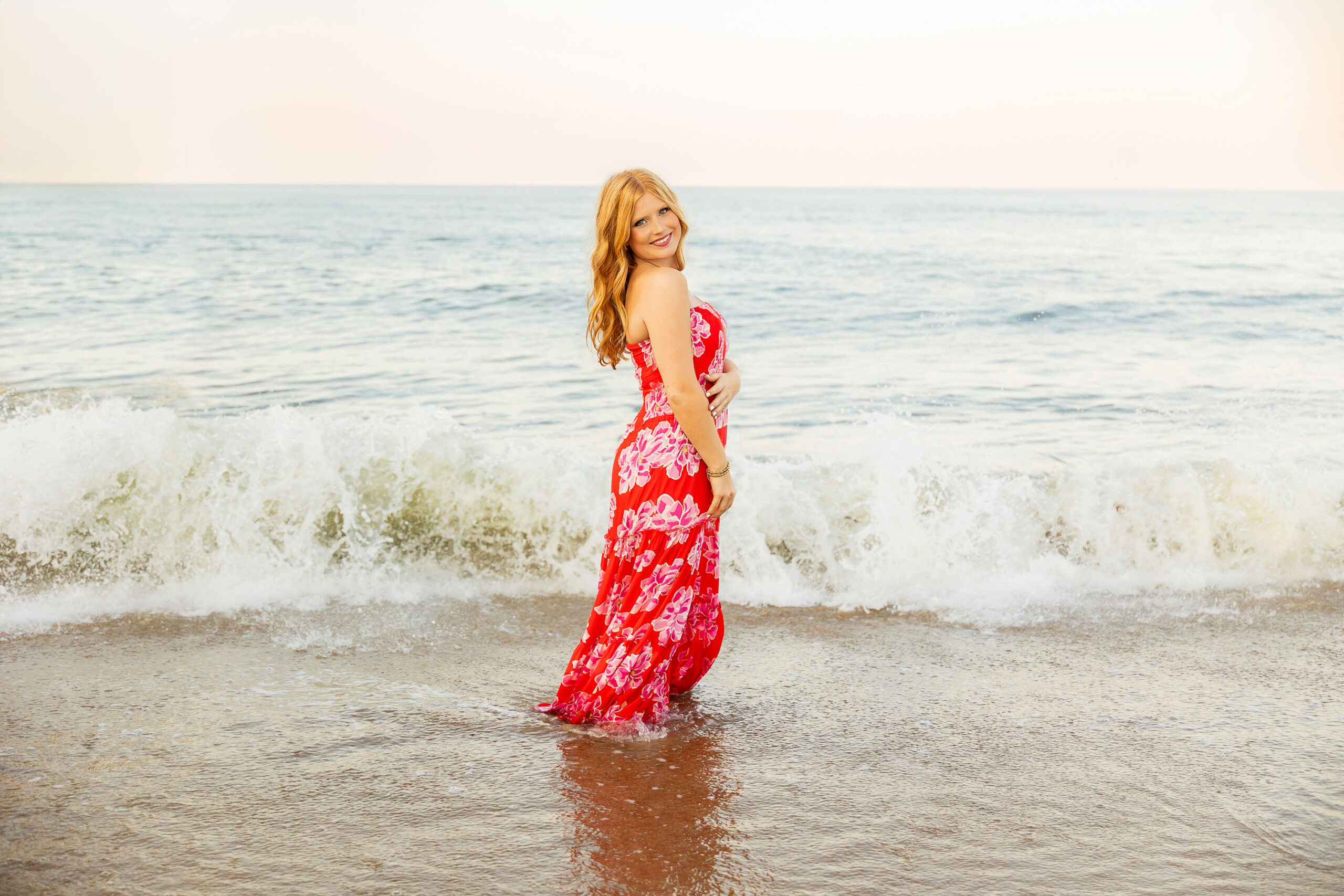 Senior girl laying in the shallow ocean waves wearing an orange floral dress during sunset at Kitty Hawk Fishing Pier.