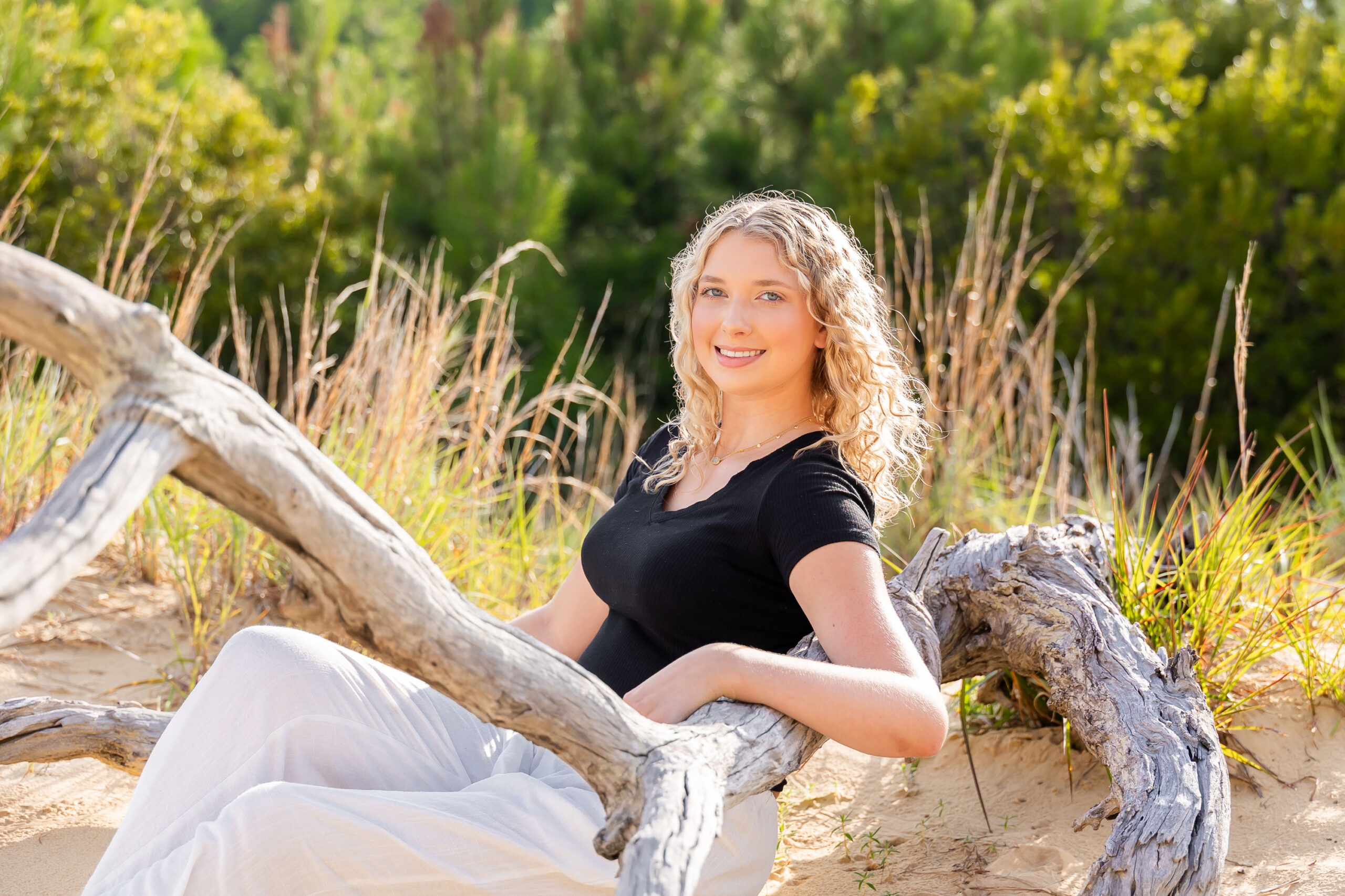 Lia posing among the sand dunes and sea oats at Jockey's Ridge, wearing a black top and white pants, with tall grasses and driftwood around her.