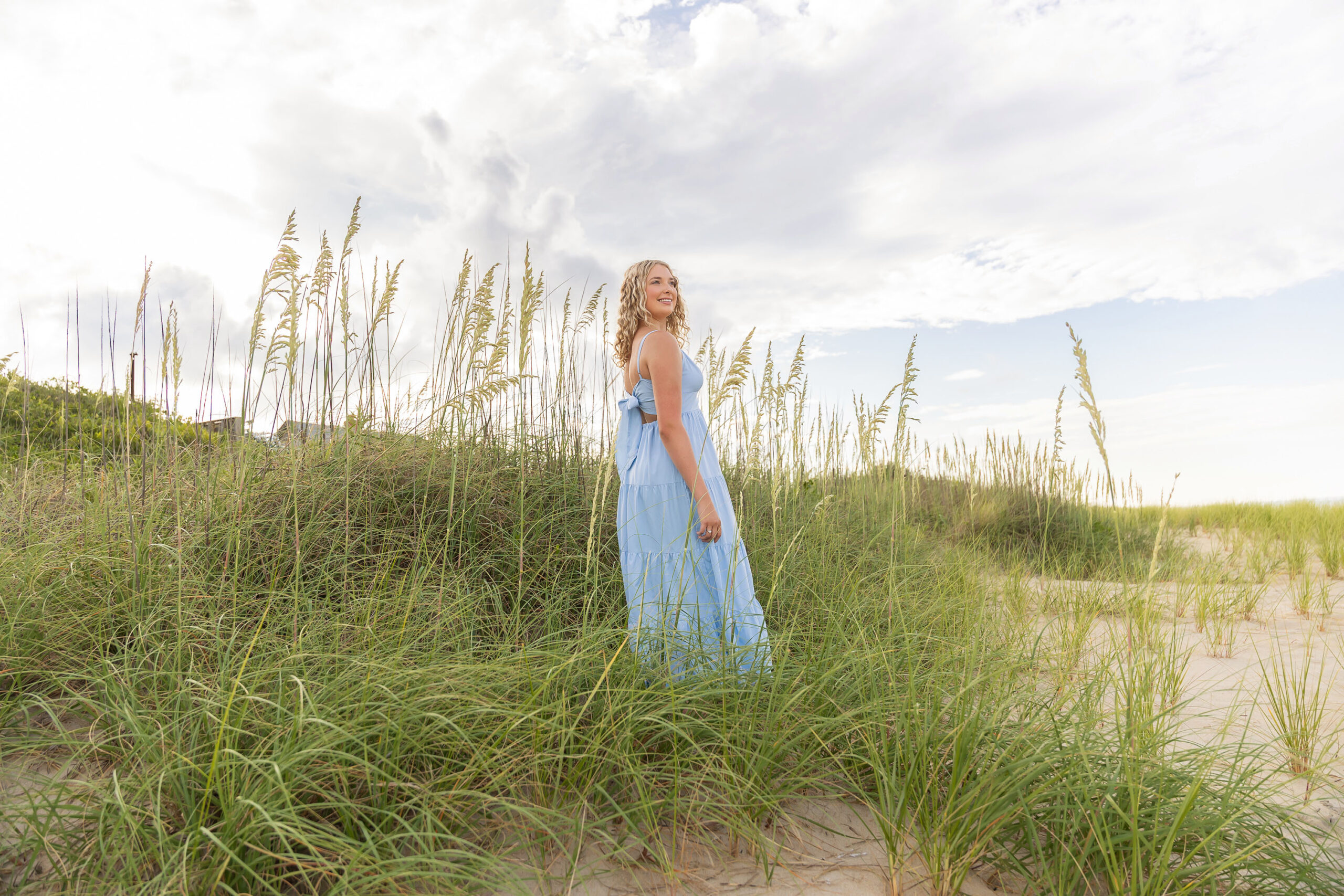 Lia in a flowing light blue dress walking along the sandy Outer Banks beach at sunset, with a soft golden glow.