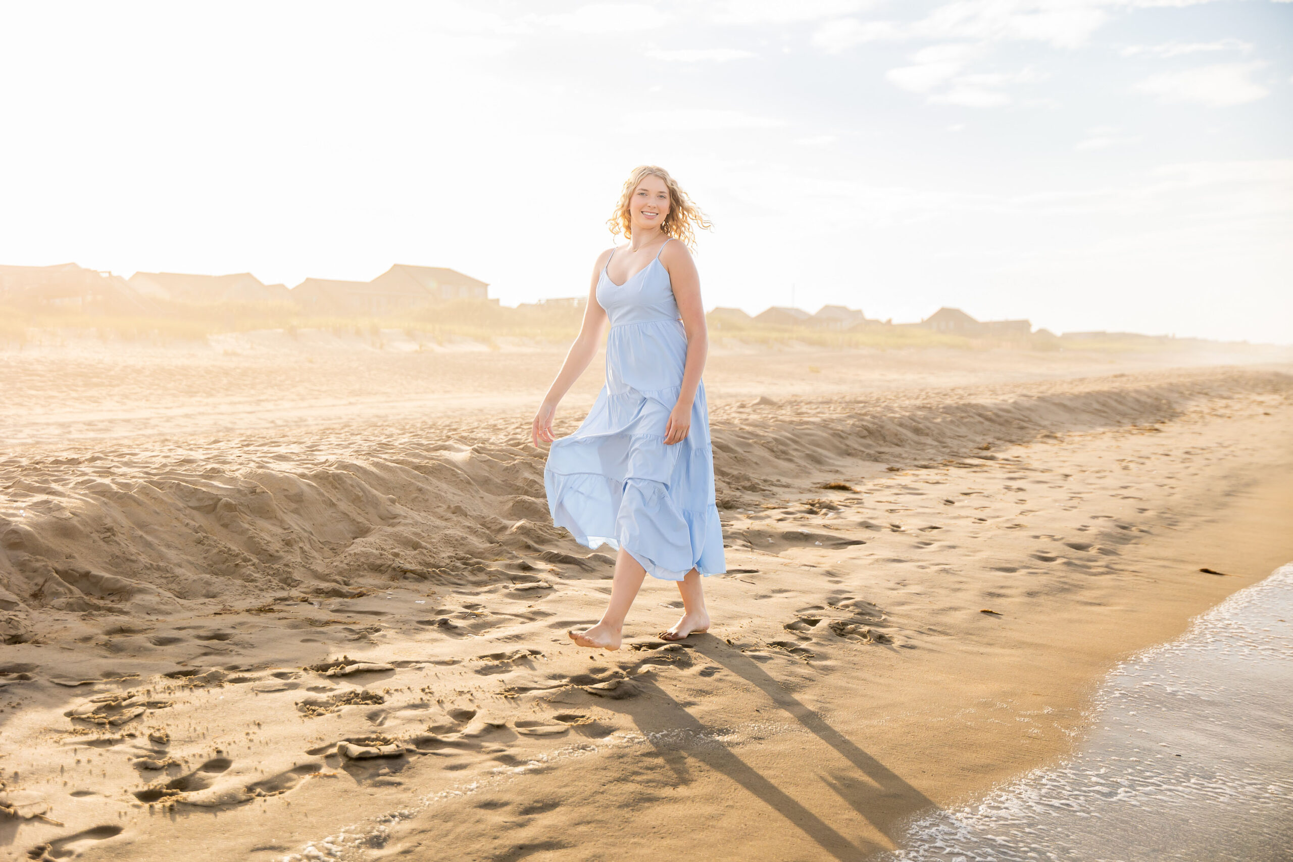 Lia in a flowing light blue dress walking along the sandy Outer Banks beach at sunset, with a soft golden glow.