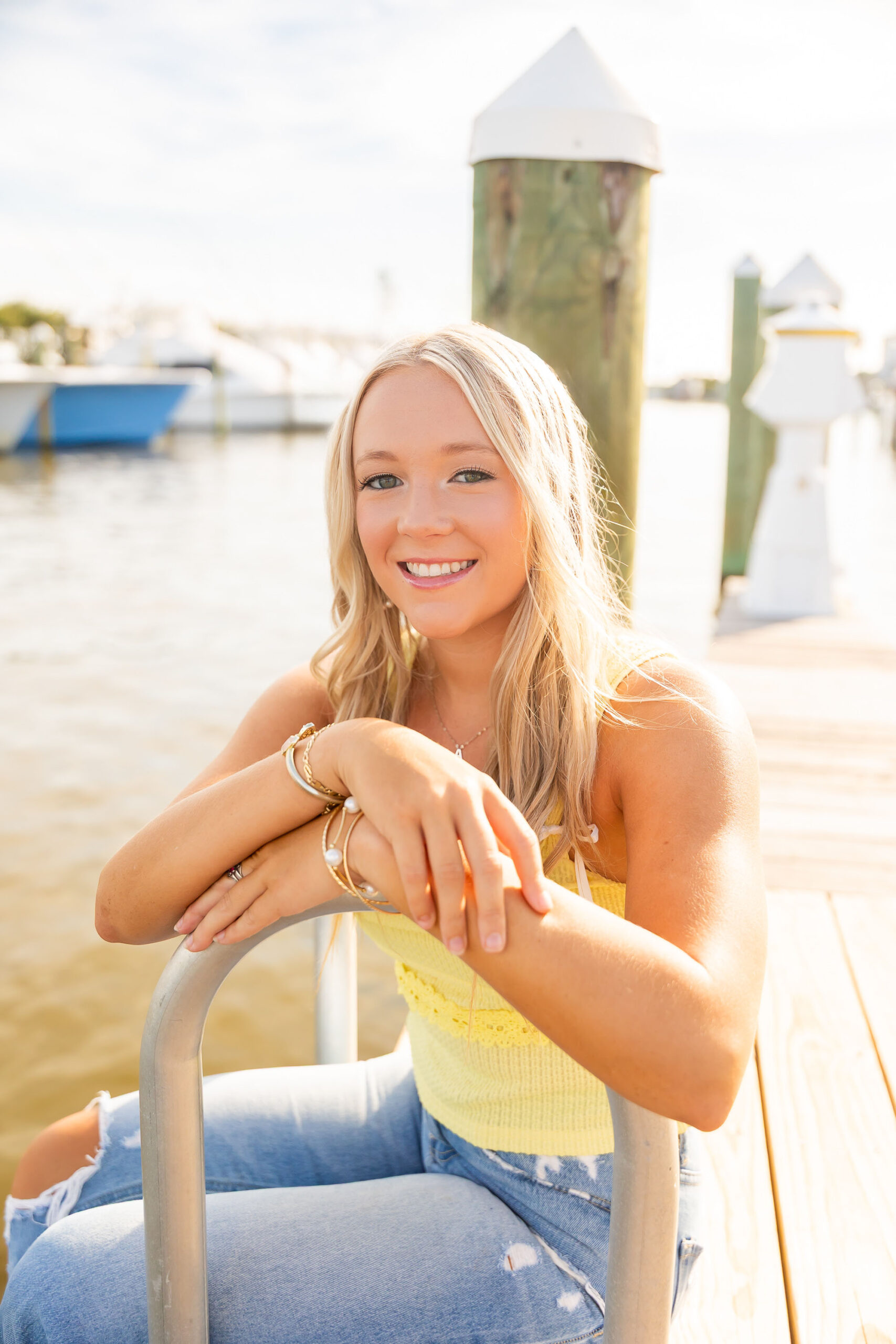 High school senior Annabelle sitting on the dock at Pirate’s Cove Marina, wearing a bright yellow top and jeans, smiling with boats in the background.