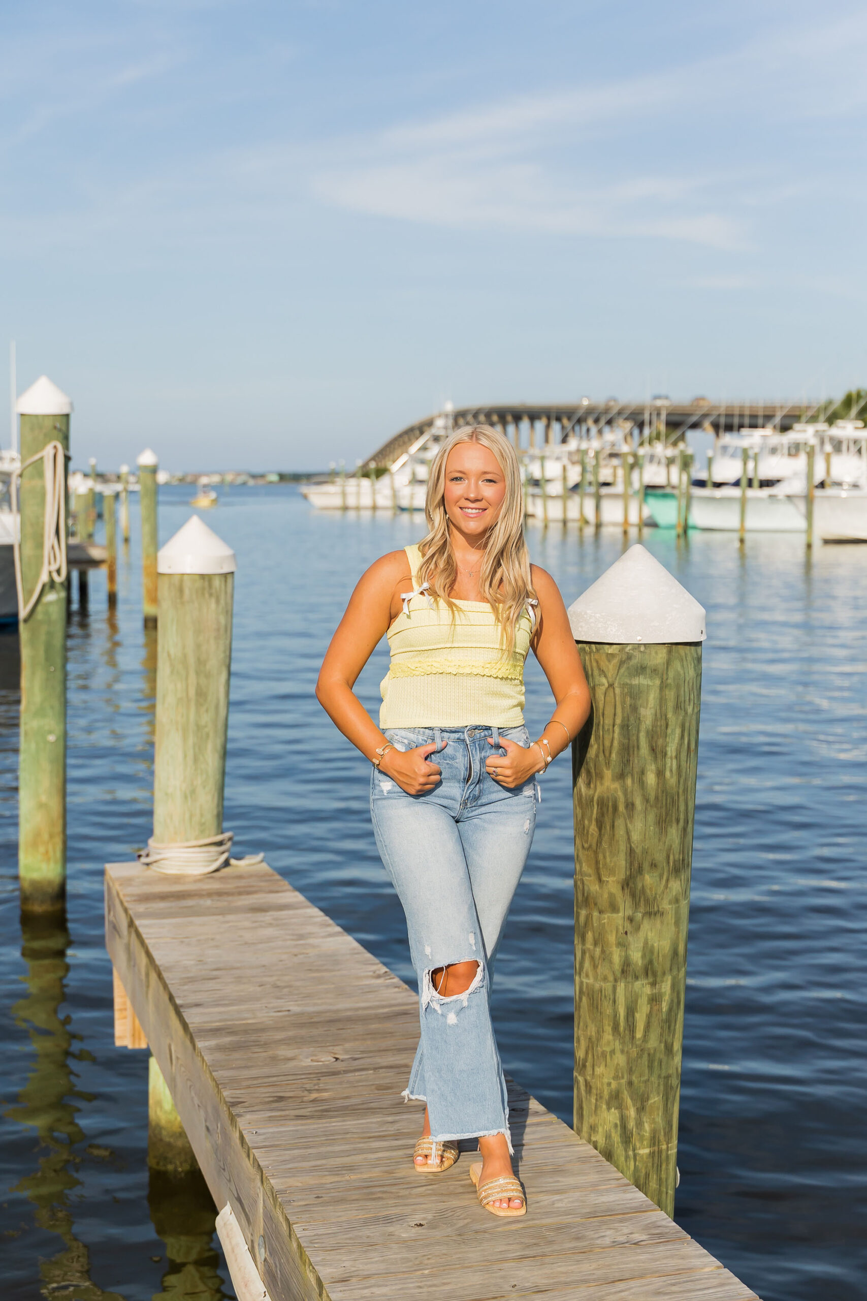 High school senior Annabelle sitting on the dock at Pirate’s Cove Marina, wearing a bright yellow top and jeans, smiling with boats in the background.