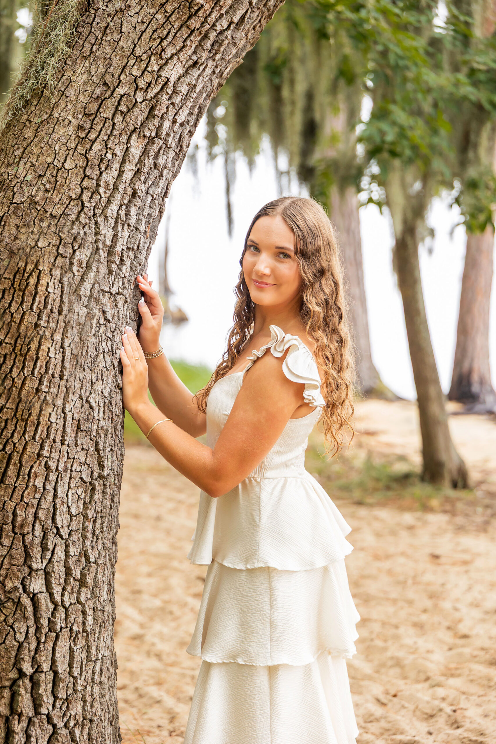Lilyanne barefoot under Spanish moss trees at Goose Creek State Park, posing in a tiered white dress during her senior portrait session.