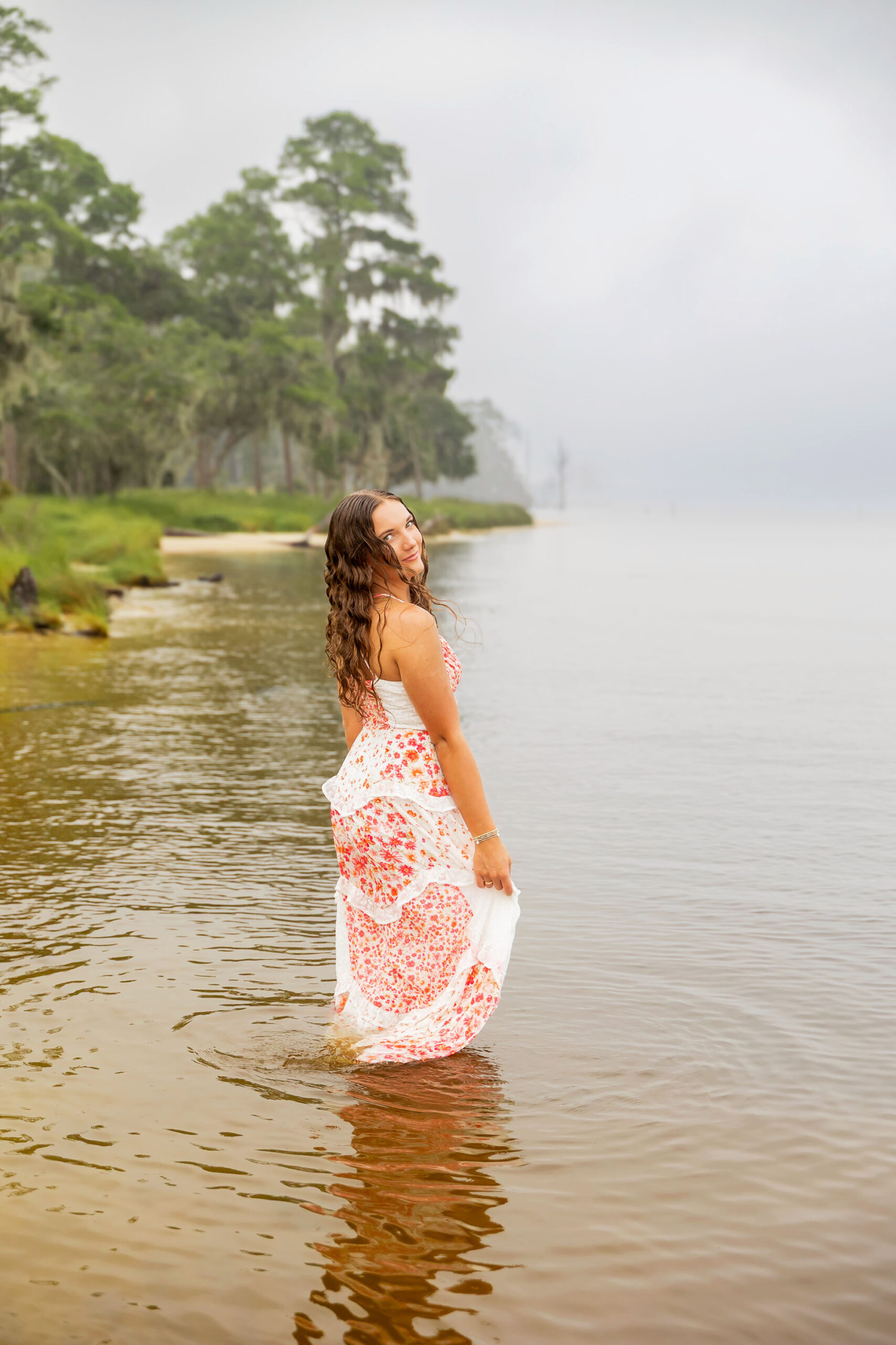 Lilyanne standing in the water at Goose Creek State Park during her senior portrait session, wearing a floral dress and embracing the misty weather.