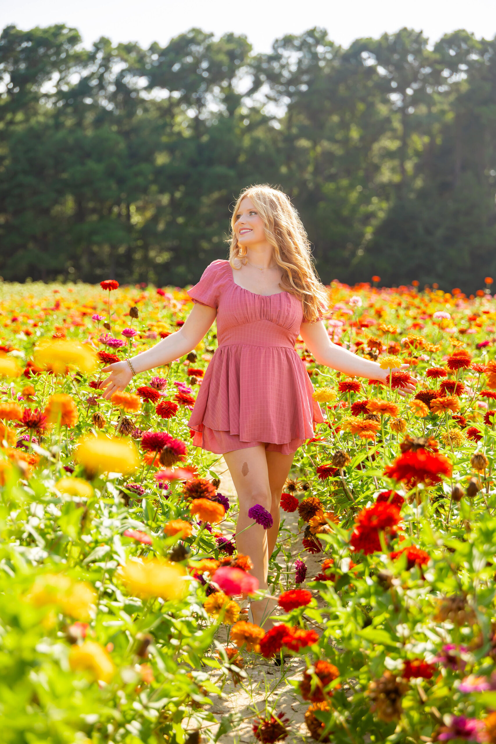 Senior girl standing in a wildflower field wearing a pink dress, surrounded by soft pink and green blooms in natural light.