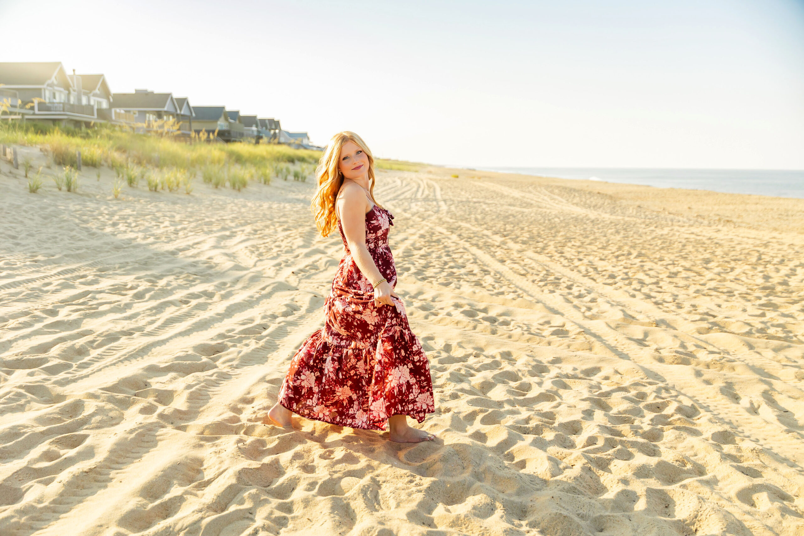 Senior girl standing in the beach dunes wearing a maroon floral maxi dress with sea grass and coastline behind her.