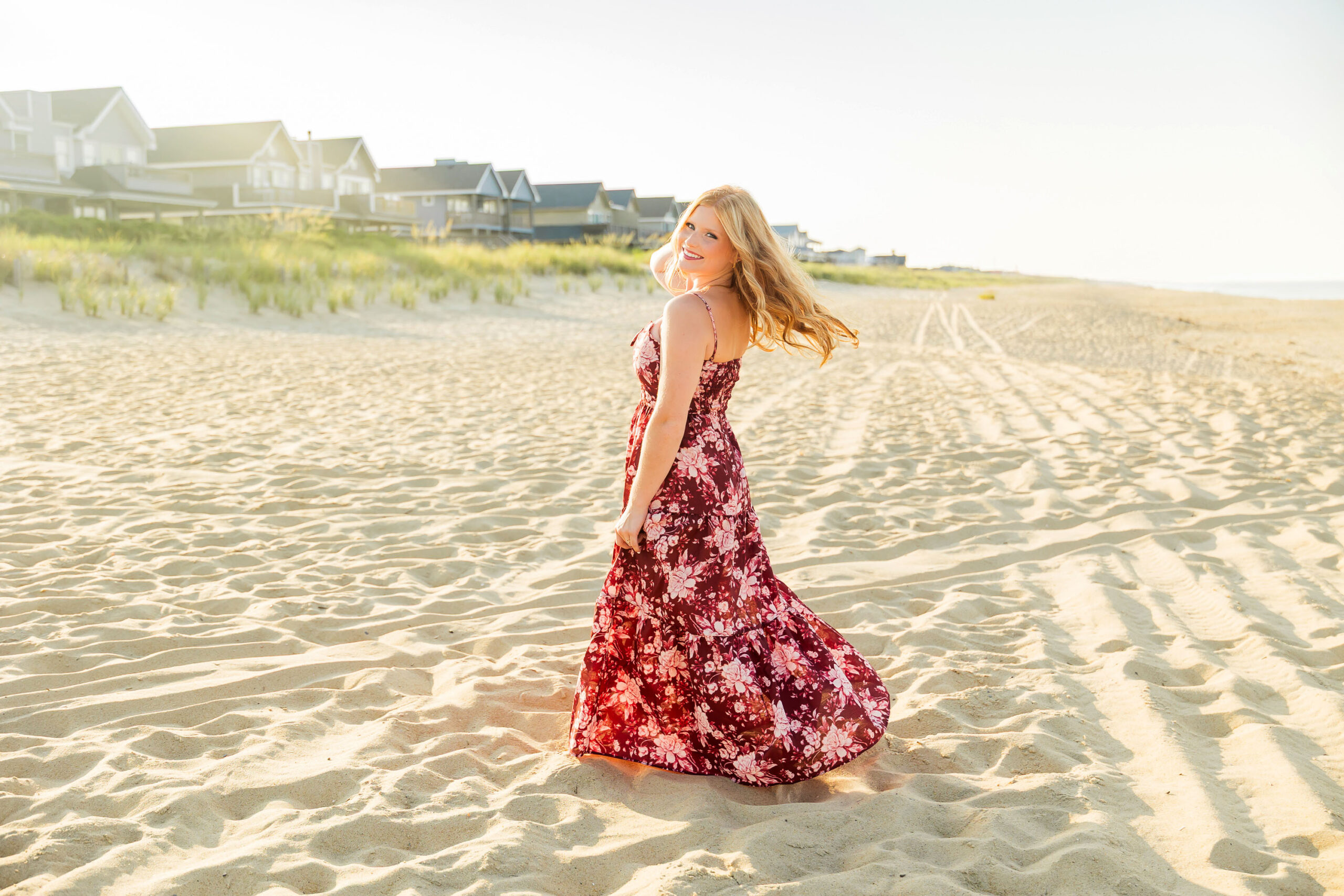 Senior girl standing in the beach dunes wearing a maroon floral maxi dress with sea grass and coastline behind her.