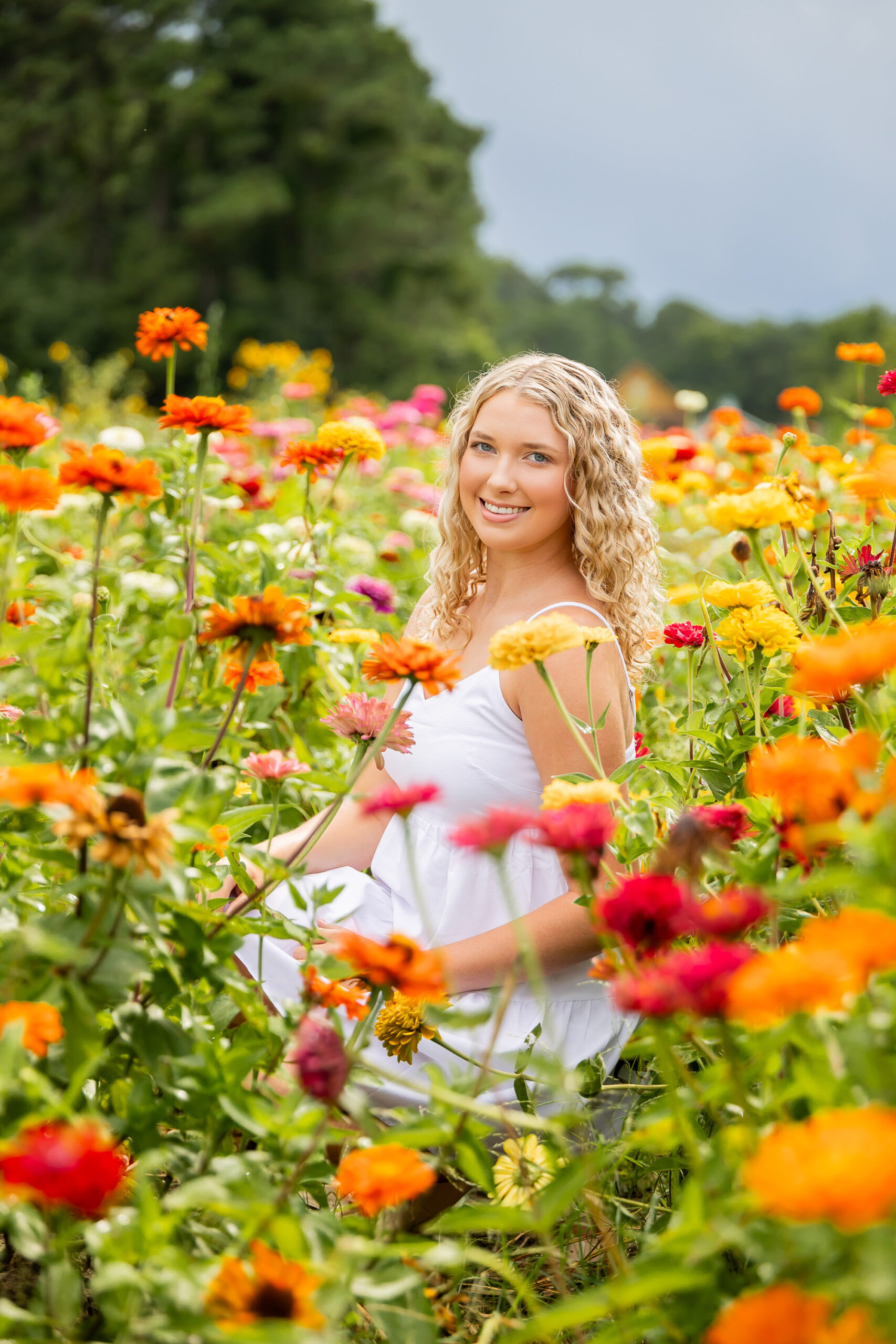 Lia sitting in a vibrant field of zinnias and sunflowers at Grandy Greenhouse, with colorful blooms all around her and a hint of storm clouds in the sky.