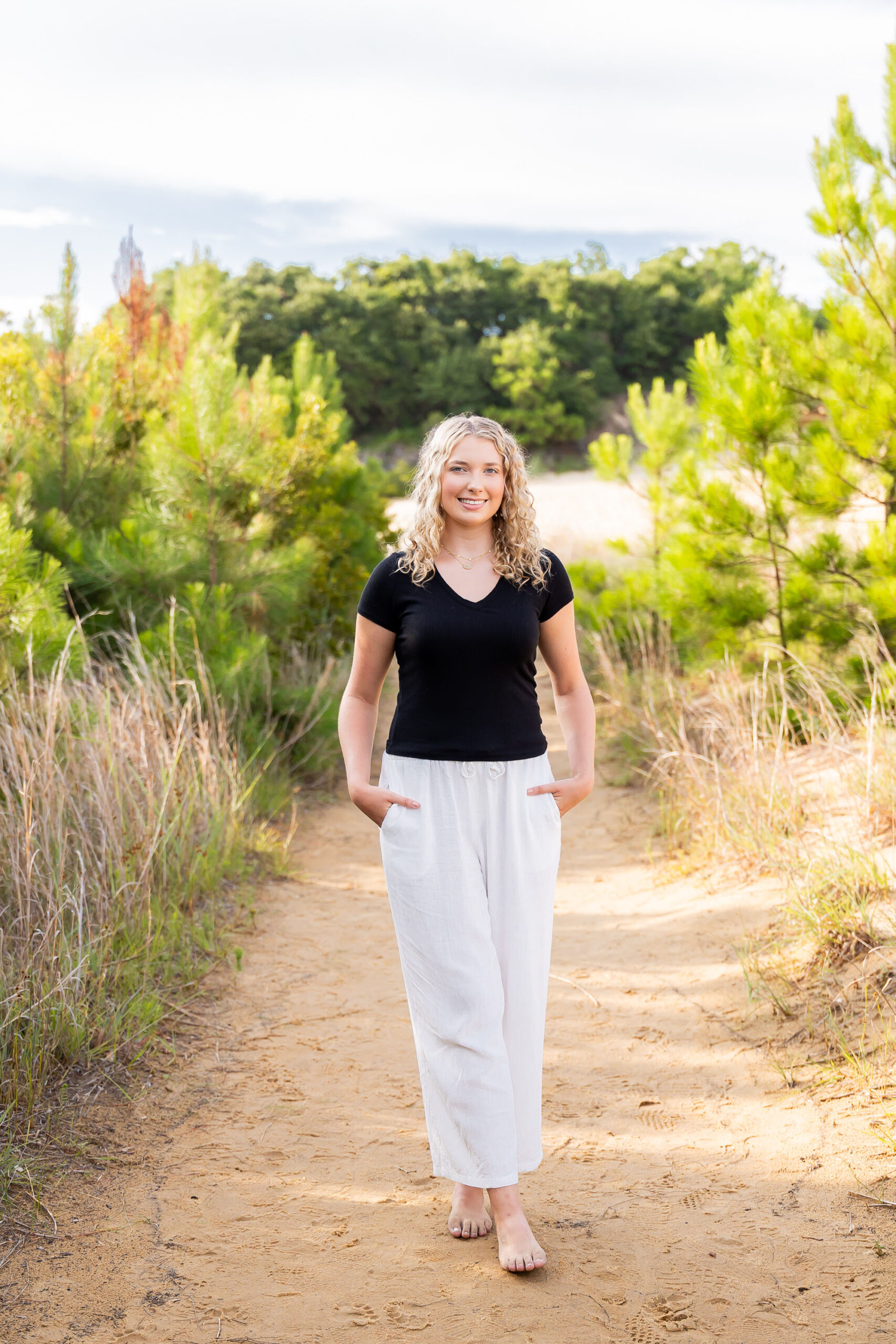 Lia posing among the sand dunes and sea oats at Jockey's Ridge, wearing a black top and white pants, with tall grasses and driftwood around her.