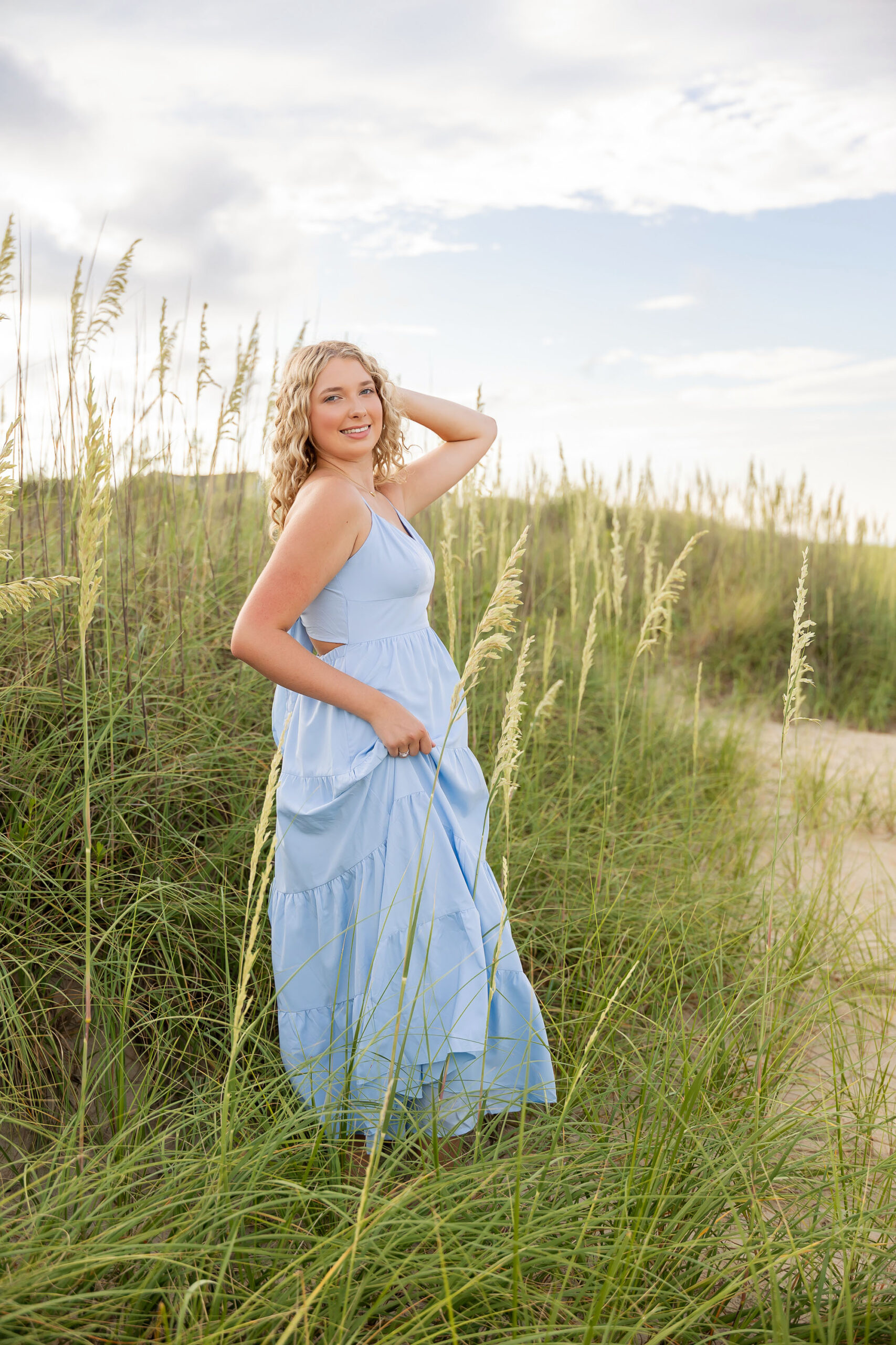 Lia in a flowing light blue dress walking along the sandy Outer Banks beach at sunset, with a soft golden glow.