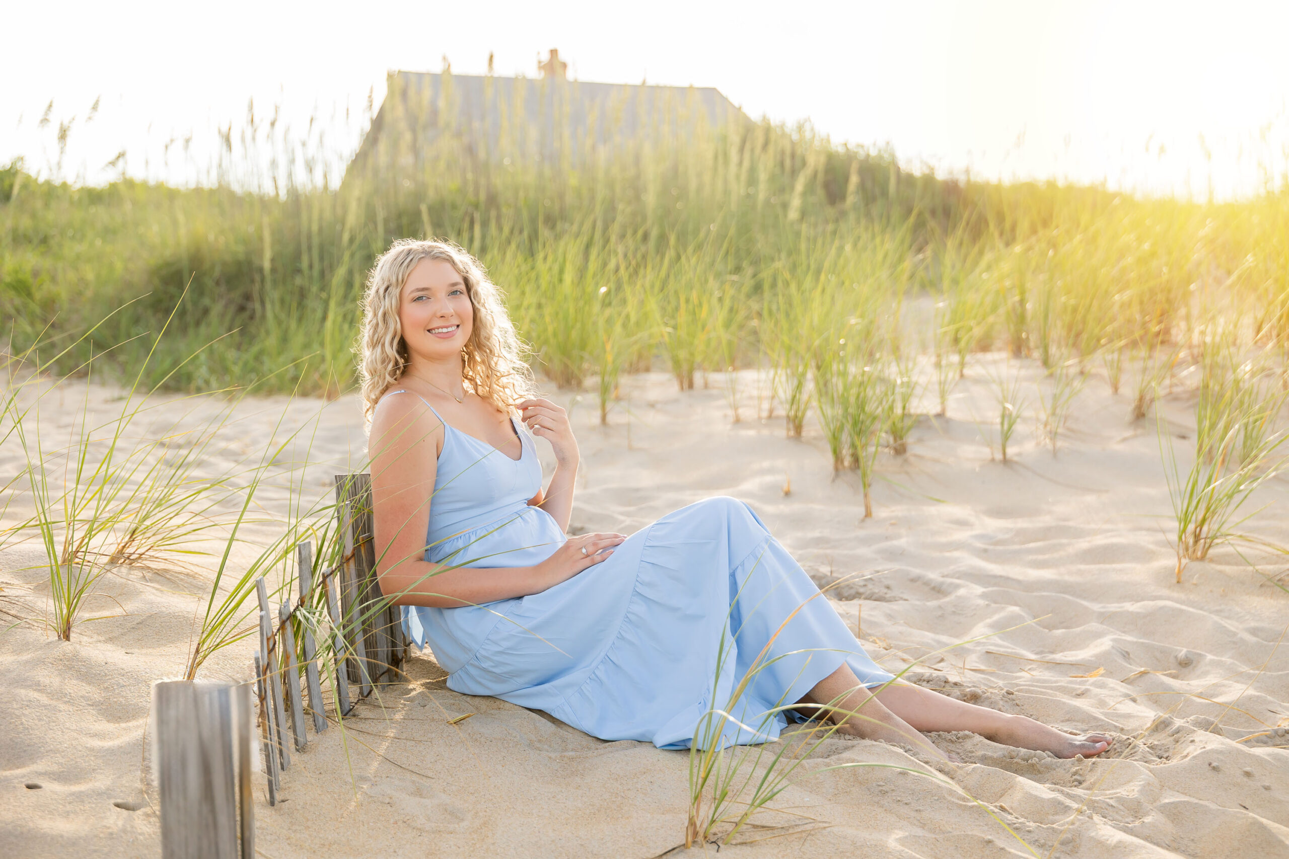 Lia in a flowing light blue dress walking along the sandy Outer Banks beach at sunset, with a soft golden glow.
