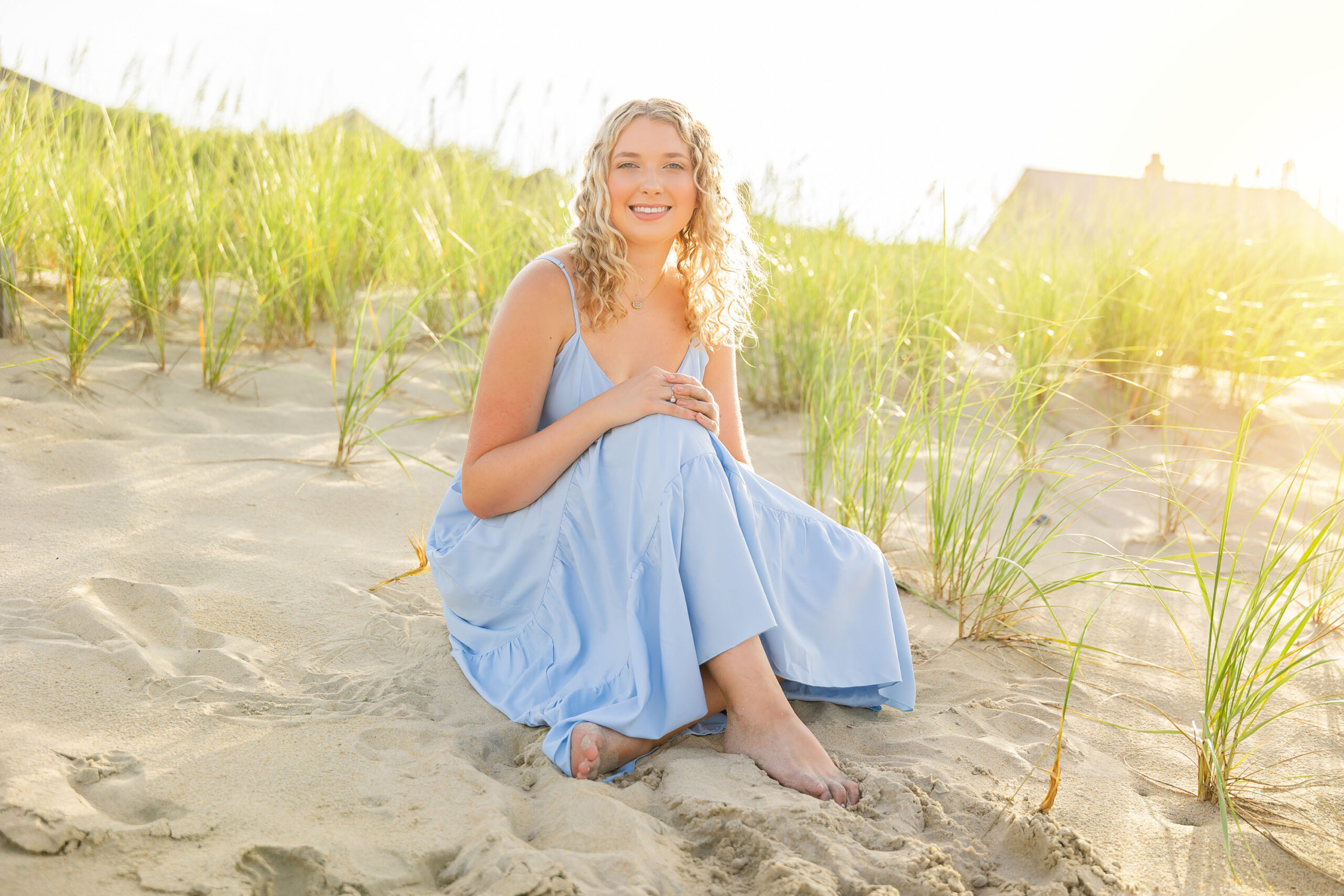 Lia in a flowing light blue dress walking along the sandy Outer Banks beach at sunset, with a soft golden glow.