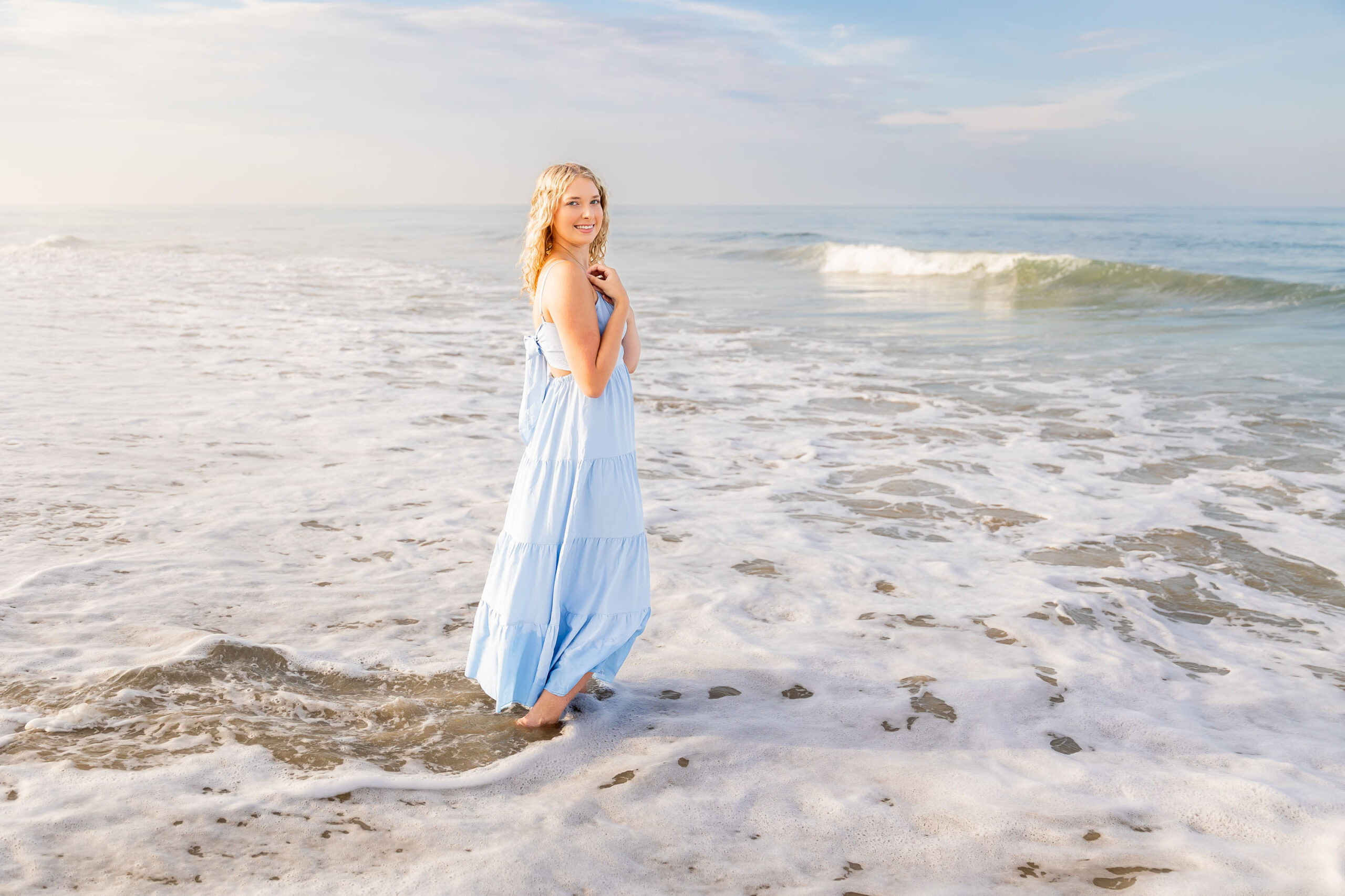 Lia in a flowing light blue dress walking along the sandy Outer Banks beach at sunset, with a soft golden glow.