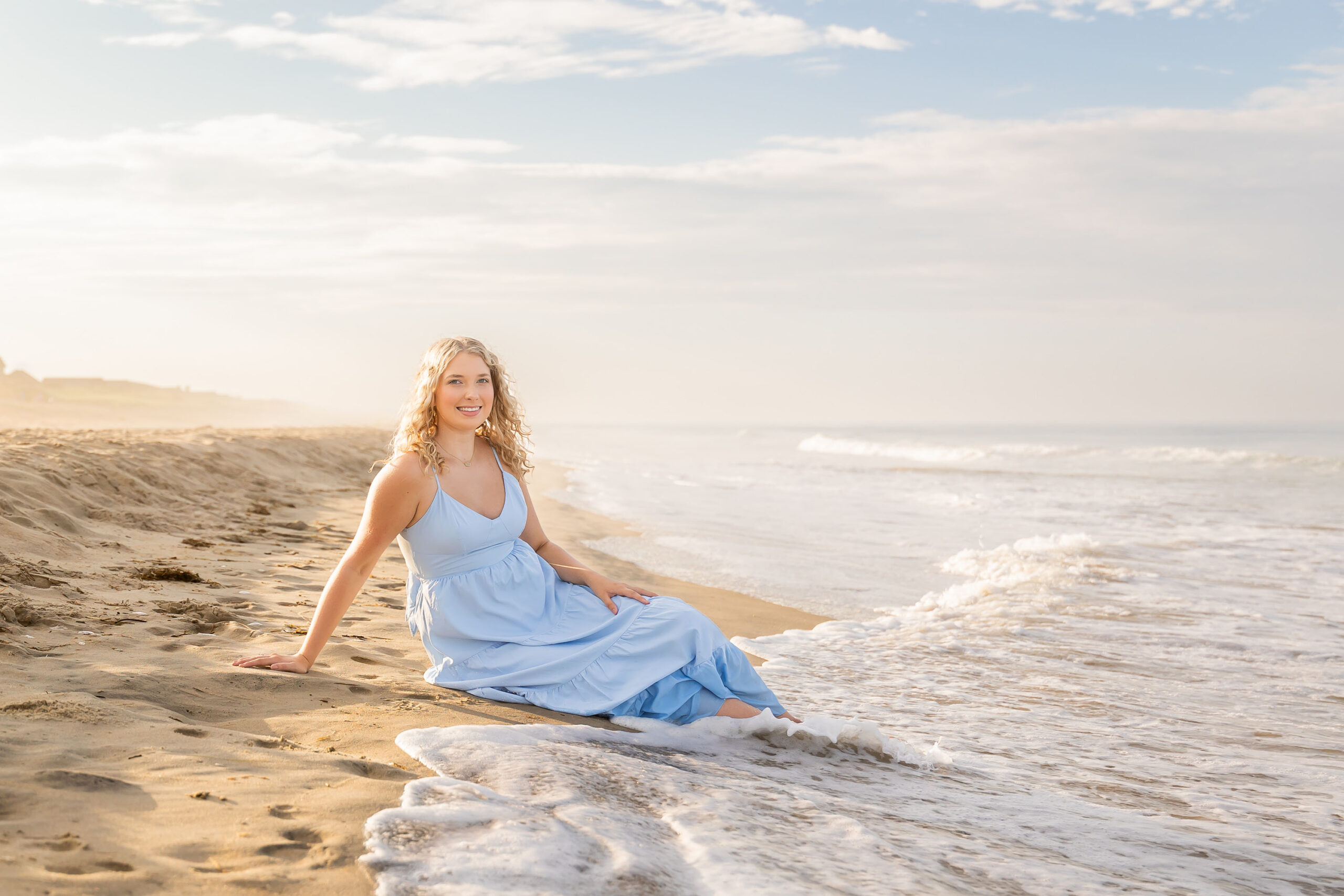 Lia in a flowing light blue dress walking along the sandy Outer Banks beach at sunset, with a soft golden glow.
